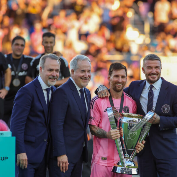 Lionel Messi and David Beckham holding the 2025 MLS Cup on a stage surrounded by Inter Miami players and personnel.